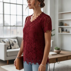 Puede incluir: Una blusa de encaje color burdeos oscuro de manga corta con escote y dobladillo festoneados. La prenda presenta un intrincado patrón de encaje floral en toda su superficie, lo que le confiere un aspecto texturizado. Se combina con pantalones vaqueros azul claro.