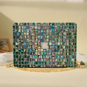 May include: A laptop hard shell case featuring a grid of small, square tiles in shades of iridescent green, teal, and silver. The surface has a reflective, metallic finish with a central cutout revealing the silver Apple logo on the laptop lid.