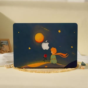 May include: A dark blue laptop skin featuring a graphic illustration of a small figure with orange hair sitting on a planet, looking at a bright yellow sun and a red rose. The white Apple logo is visible in the center of the starry night sky.