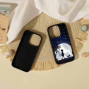 May include: Two black smartphone cases. One case is plain black, while the other features a graphic illustration of a silhouette of a person and a cat walking under a large white moon against a dark blue starry night sky with twisted trees.