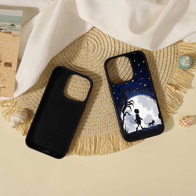 May include: Two black smartphone cases. One case is plain black, while the other features a graphic illustration of a silhouette of a person and a cat walking under a large white moon against a dark blue starry night sky with twisted trees.