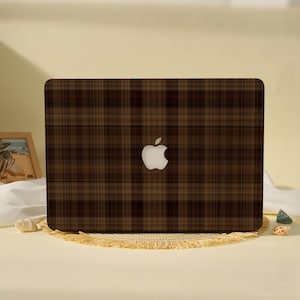 May include: A rectangular laptop skin featuring a dark brown and black plaid pattern with a centered cutout for the white Apple logo. The design consists of intersecting horizontal and vertical lines creating a classic grid motif across the surface.