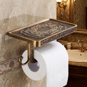 May include: An antique-style toilet paper holder with a decorative shelf. The holder is made of bronze-colored metal and features ornate scrollwork and a detailed design on the shelf. A roll of white toilet paper is in place.