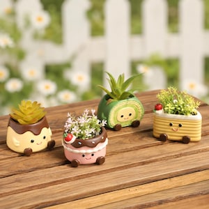 Pode incluir: Quatro pequenos vasos de cerâmica em forma de sobremesas, incluindo um pudim, um bolo rosa, um rocambole verde e um bolo retangular amarelo. Cada vaso apresenta um rosto sorridente, pequenos pés castanhos e uma planta artificial.