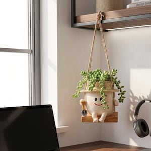 May include: A white ceramic planter shaped like a smiling character wearing a straw hat, holding a small shovel, and sitting on a wooden swing. The planter is suspended by a rope and contains a trailing green succulent plant with round leaves.