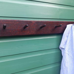 Pode incluir: Um cabide de parede retangular de madeira castanho-escura com quatro ganchos salientes espaçados uniformemente. Uma camisa às riscas azul-claras e brancas e uma peça de vestuário cor de vinho lisa estão penduradas nos dois ganchos da direita.