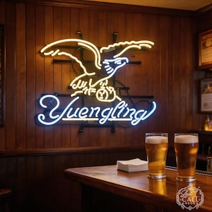 May include: A neon sign featuring a white eagle design with the word "Yuengling" in blue neon. Two glasses of beer sit on a wooden bar. The sign is mounted on a wood-paneled wall.