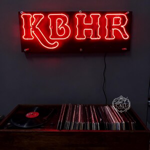Könnte beinhalten: Ein rechteckiges Neonschild mit den Buchstaben KBHR in hellem Rotlicht, montiert auf einer dunklen rechteckigen Rückwand. Unter dem Schild befindet sich eine hölzerne Plattenspielerkonsole mit einer schwarzen Schallplatte und einer Plattensammlung.