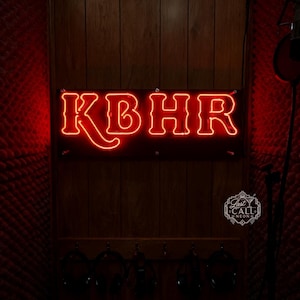 Könnte beinhalten: Ein rechteckiges Neonschild mit den Buchstaben KBHR in leuchtendem Rot, montiert an einer holzverkleideten Wand. Unter dem Schild hängen fünf schwarze Kopfhörer an Haken an einer Holzleiste. Ein kleines weißes Logo ist unten rechts zu sehen.