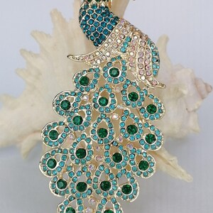 May include: A gold-toned peacock brooch with a detailed design. The peacock's body and tail feathers are adorned with teal, green, and pink rhinestones. The brooch is set against a white background, highlighting its intricate details.