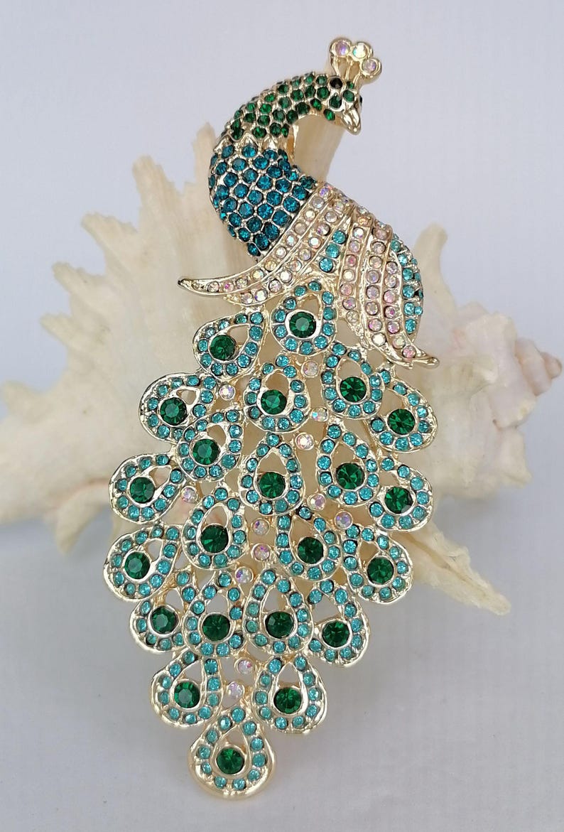 May include: A gold-toned peacock brooch with a detailed design. The peacock's body and tail feathers are adorned with teal, green, and pink rhinestones. The brooch is set against a white background, highlighting its intricate details.