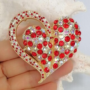 May include: A heart-shaped brooch with a gold-tone metal frame. The brooch is adorned with red and iridescent crystals. The outer heart is outlined with smaller red crystals. A romantic accessory.