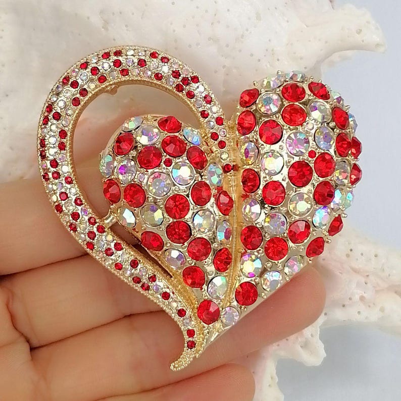 May include: A heart-shaped brooch with a gold-tone metal frame. The brooch is adorned with red and iridescent crystals. The outer heart is outlined with smaller red crystals. A romantic accessory.