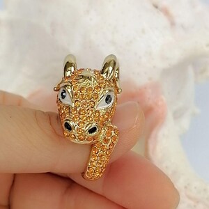 May include: A gold-toned ring featuring a three-dimensional ram head design. The ram is covered in small, sparkling orange crystals, with smooth white enamel around the eyes and black accents for the pupils and nostrils, set on a gold band.