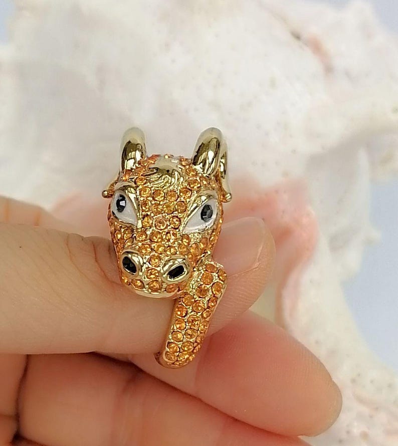 May include: A gold-toned ring featuring a three-dimensional ram head design. The ram is covered in small, sparkling orange crystals, with smooth white enamel around the eyes and black accents for the pupils and nostrils, set on a gold band.