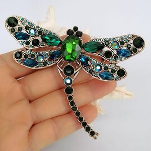 May include: A gold-coloured dragonfly brooch, embellished with emerald green and sapphire blue crystals. The wings are decorated with round and marquise-cut crystals. The body features a large, central green crystal and a tail of smaller green stones.