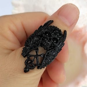 以下が含まれることがあります： 小さな黒いクリスタルで飾られた中央の頭蓋骨モチーフが特徴の黒い指輪。頭蓋骨の両側には質感のある翼があり、その下には交差した骨が配置されています。全体はダークでマットな黒い金属調の仕上げになっています。