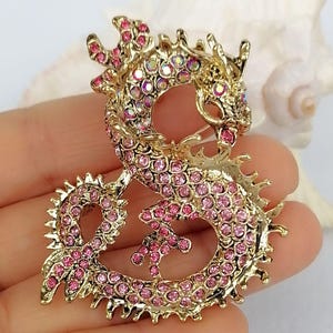 Puede incluir: Broche de drag&oacute;n dorado con brillantes rosas. El drag&oacute;n est&aacute; en una pose estilizada, con su cuerpo formando un bucle. El broche tiene un alfiler en la parte posterior para una fijaci&oacute;n segura. Las escamas y los detalles del drag&oacute;n est&aacute;n claramente definidos.
