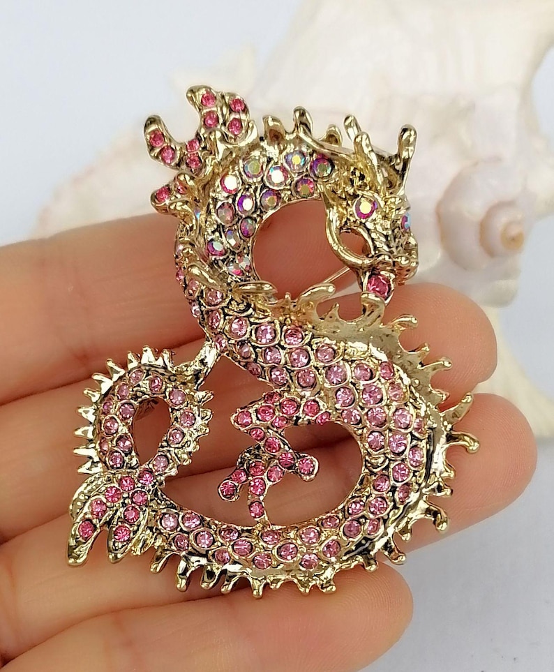 Puede incluir: Broche de drag&oacute;n dorado con brillantes rosas. El drag&oacute;n est&aacute; en una pose estilizada, con su cuerpo formando un bucle. El broche tiene un alfiler en la parte posterior para una fijaci&oacute;n segura. Las escamas y los detalles del drag&oacute;n est&aacute;n claramente definidos.