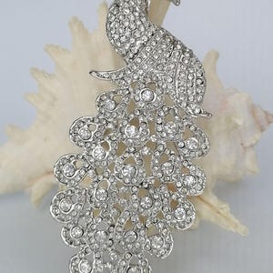 May include: A silver-toned peacock brooch, encrusted with numerous clear rhinestones. The brooch is in the shape of a peacock, with its tail feathers fanned out. The brooch is set against a white background.