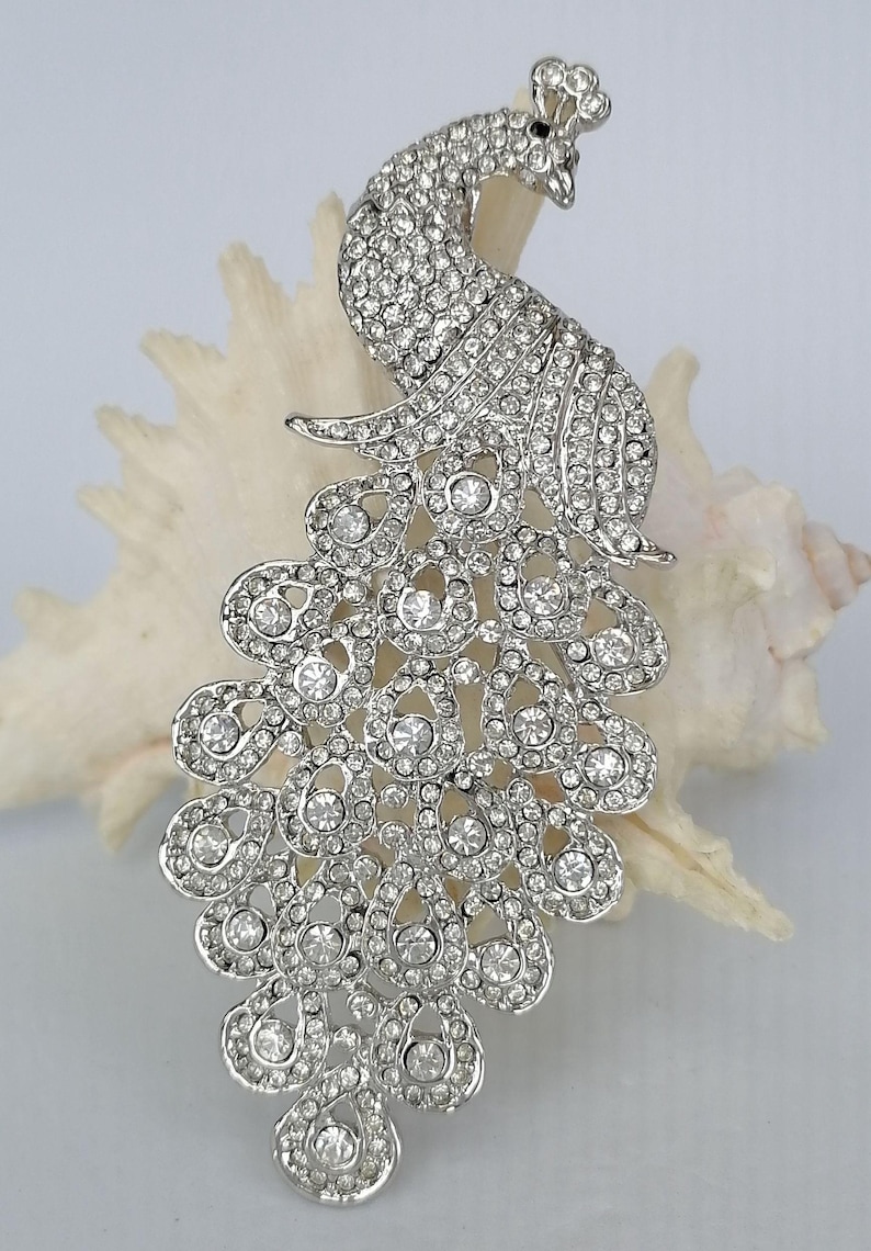 May include: A silver-toned peacock brooch, encrusted with numerous clear rhinestones. The brooch is in the shape of a peacock, with its tail feathers fanned out. The brooch is set against a white background.