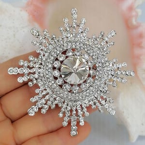 K&ouml;nnte beinhalten: Eine silberfarbene Brosche in Schneeflockenform mit einem gro&szlig;en, facettierten, klaren Mittelstein, der von mehreren konzentrischen Kreisen aus kleineren, funkelnden, klaren Strasssteinen umgeben ist. Das komplexe Sternendesign strahlt mit kristallbesetzten Spitzen.