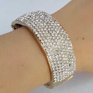 May include: A wide gold-tone hinged bangle bracelet featuring a dense, multi-row pavé setting of clear, sparkling round rhinestones covering the entire outer surface. The metal frame has a polished gold finish visible along the edges.