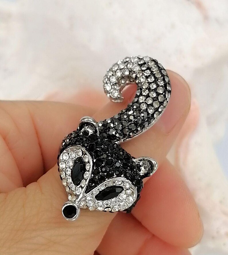 K&ouml;nnte beinhalten: Eine Brosche in Form eines Stinktierkopfes mit geschwungenem Schwanz, bestehend aus einer silberfarbenen Metallbasis, die mit zahlreichen kleinen schwarzen und klaren Kristallen besetzt ist, sowie zwei gro&szlig;en schwarzen ovalen Steinen als Augen.