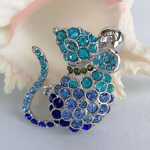 May include: A silver-toned mouse brooch adorned with blue and teal rhinestones. The brooch features a rounded body, a curved tail, and a large, round head. The rhinestones are arranged in a clustered pattern, creating a sparkling effect.