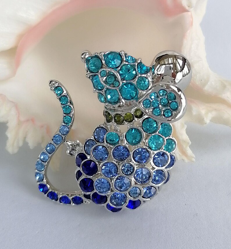 May include: A silver-toned mouse brooch adorned with blue and teal rhinestones. The brooch features a rounded body, a curved tail, and a large, round head. The rhinestones are arranged in a clustered pattern, creating a sparkling effect.