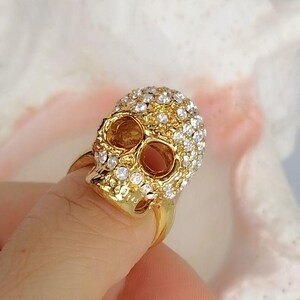Op de afbeelding: Een goudkleurige ring met een driedimensionaal schedelontwerp. Het bovenste gedeelte van de schedel is ingelegd met talloze kleine, heldere, sprankelende strass-steentjes, terwijl de onderkaak en oogkassen een gladde, gepolijste gouden afwerking hebben.