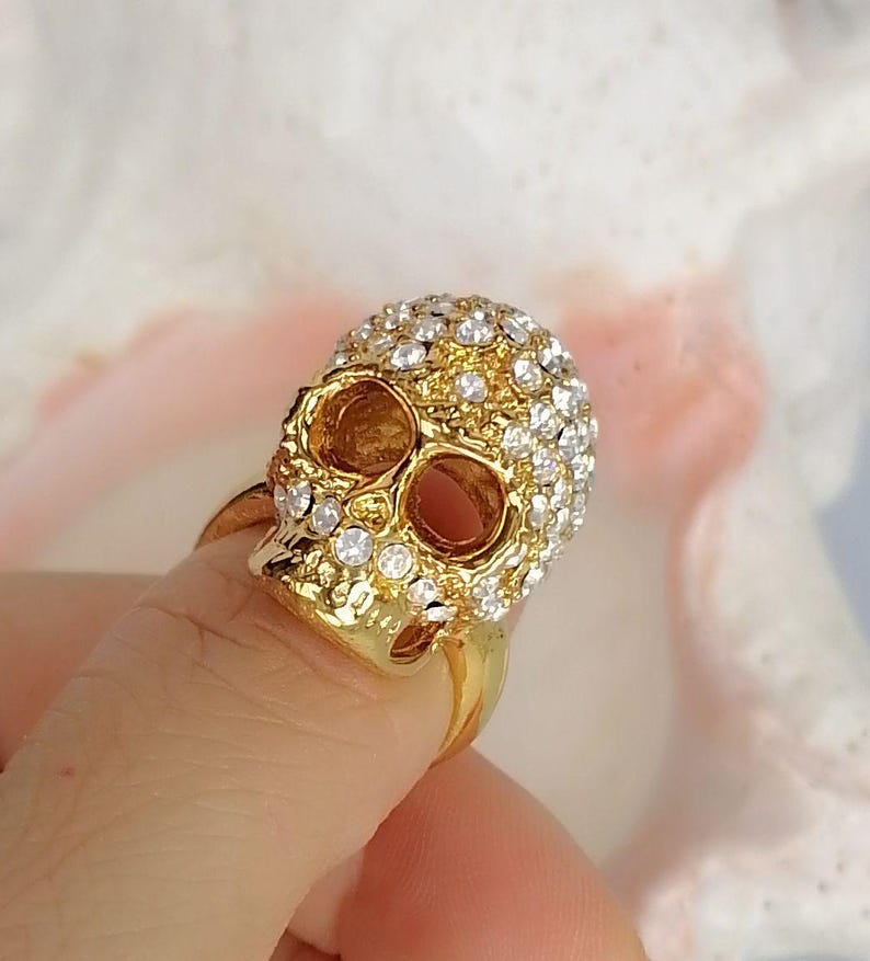 Op de afbeelding: Een goudkleurige ring met een driedimensionaal schedelontwerp. Het bovenste gedeelte van de schedel is ingelegd met talloze kleine, heldere, sprankelende strass-steentjes, terwijl de onderkaak en oogkassen een gladde, gepolijste gouden afwerking hebben.