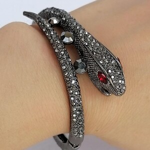 Op de afbeelding: Een slangvormige armband van donkerkleurig metaal, volledig ingelegd met kleine, glinsterende zwarte kristallen. De slangenkop heeft twee felrode kristallen ogen en het lichaam is geaccentueerd met drie grotere ronde zwarte stenen.