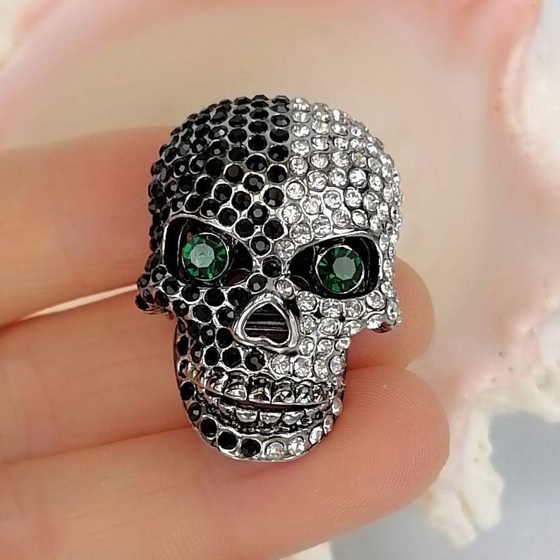 Pode incluir: Um broche em forma de caveira com um design dividido coberto por pequenos cristais brilhantes. O lado esquerdo &eacute; adornado com cristais pretos, enquanto o lado direito apresenta cristais transparentes. Dois cristais verdes vibrantes servem como olhos.
