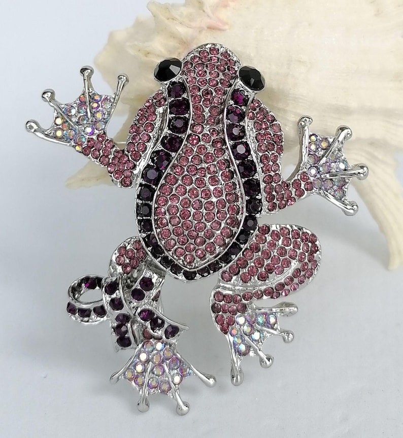 K&ouml;nnte beinhalten: Silberfarbene Froschbrosche mit rosa und lila Kristallen. Der Froschk&ouml;rper ist mit kleinen, runden Kristallen bedeckt, mit schwarzen Kristallen f&uuml;r die Augen. Die Brosche hat ausgestreckte Arme und Beine, vor wei&szlig;em Hintergrund.
