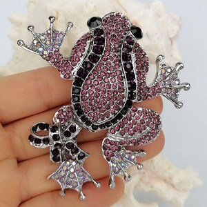 K&ouml;nnte beinhalten: Ein mit Juwelen besetzte Froschbrosche mit einer silberfarbenen Metallbasis. Der Frosch ist mit rosa, lila und schwarzen Strasssteinen bedeckt. Die Brosche hat ausgestreckte Arme und Beine, und die Augen sind schwarz. Ein dekoratives Accessoire.