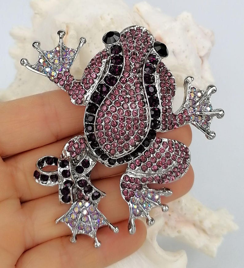 K&ouml;nnte beinhalten: Ein mit Juwelen besetzte Froschbrosche mit einer silberfarbenen Metallbasis. Der Frosch ist mit rosa, lila und schwarzen Strasssteinen bedeckt. Die Brosche hat ausgestreckte Arme und Beine, und die Augen sind schwarz. Ein dekoratives Accessoire.
