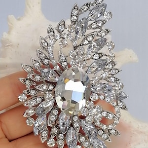 May include: A silver-toned brooch featuring a large central oval clear crystal surrounded by an intricate arrangement of smaller marquise and round clear crystals set in a leaf-like pattern. The metal frame is detailed with small pav&eacute;-set stones.