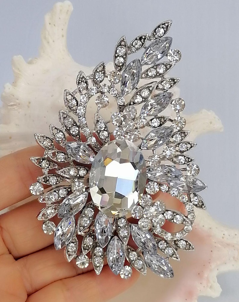 May include: A silver-toned brooch featuring a large central oval clear crystal surrounded by an intricate arrangement of smaller marquise and round clear crystals set in a leaf-like pattern. The metal frame is detailed with small pav&eacute;-set stones.