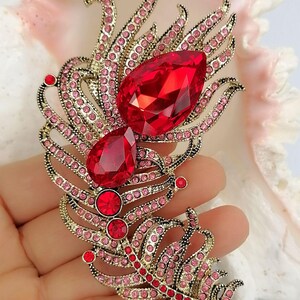May include: A gold-toned feather-shaped brooch featuring two large, faceted, teardrop-shaped red crystals, two smaller round red crystals, and numerous tiny pink rhinestones embedded along the metal frame in a decorative, intricate pattern.