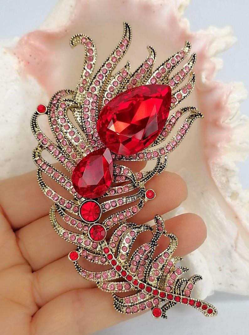 May include: A gold-toned feather-shaped brooch featuring two large, faceted, teardrop-shaped red crystals, two smaller round red crystals, and numerous tiny pink rhinestones embedded along the metal frame in a decorative, intricate pattern.
