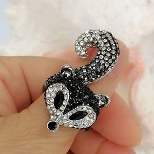 K&ouml;nnte beinhalten: Ein silberfarbener Ring in Form eines Waschb&auml;renkopfes mit geringeltem Schwanz. Das Design ist mit zahlreichen kleinen, funkelnden klaren und schwarzen Strasssteinen besetzt, mit zwei gr&ouml;&szlig;eren schwarzen Steinen als Augen und einem kleinen schwarzen Stein als Nase.