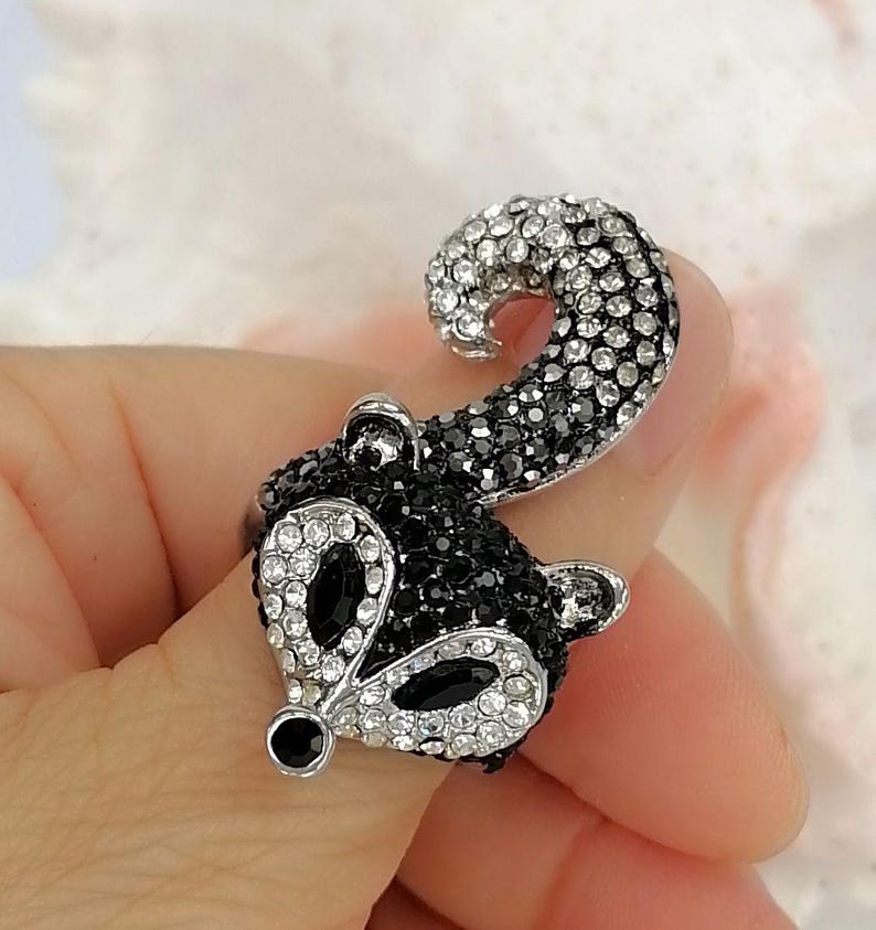 K&ouml;nnte beinhalten: Ein silberfarbener Ring in Form eines Waschb&auml;renkopfes mit geringeltem Schwanz. Das Design ist mit zahlreichen kleinen, funkelnden klaren und schwarzen Strasssteinen besetzt, mit zwei gr&ouml;&szlig;eren schwarzen Steinen als Augen und einem kleinen schwarzen Stein als Nase.