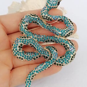 May include: A gold-coloured snake brooch adorned with teal and dark green rhinestones. The snake is coiled, with its head raised, and has a black rhinestone for an eye. The brooch is a decorative accessory.