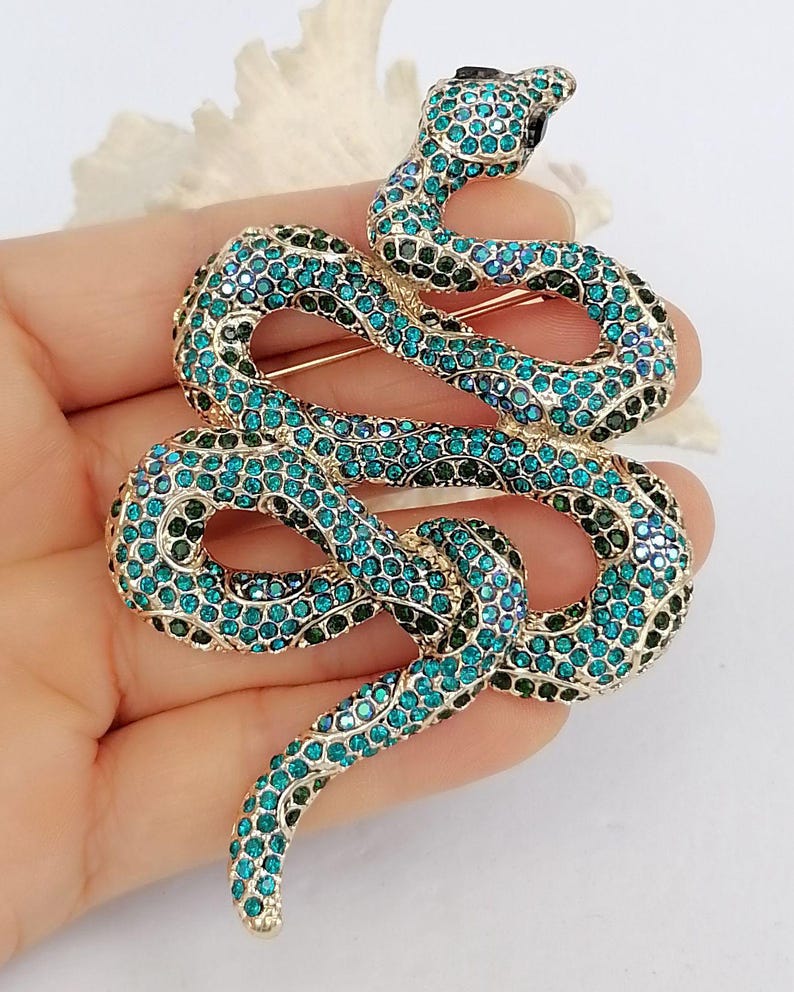 May include: A gold-coloured snake brooch adorned with teal and dark green rhinestones. The snake is coiled, with its head raised, and has a black rhinestone for an eye. The brooch is a decorative accessory.