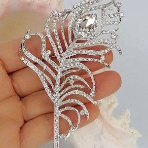 May include: Silver-coloured feather brooch, adorned with clear rhinestones. The brooch has a large, teardrop-shaped rhinestone at the centre. The design is intricate and detailed, resembling a peacock feather.