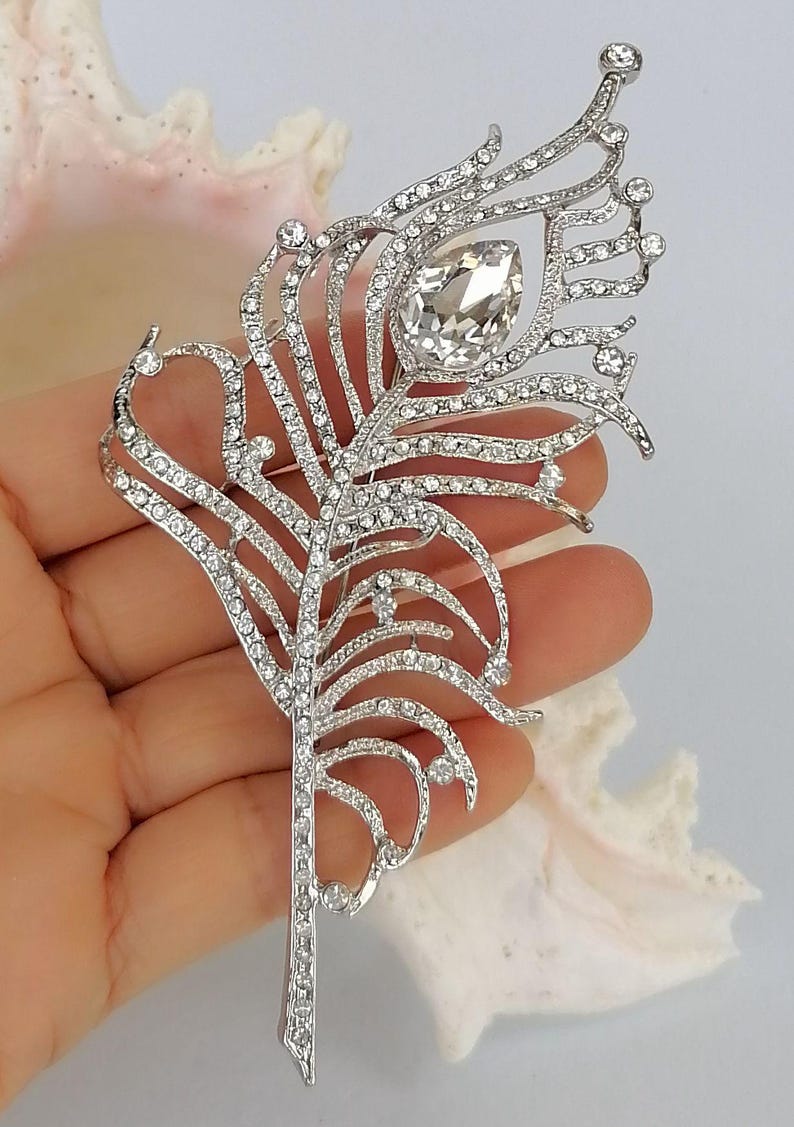 May include: Silver-coloured feather brooch, adorned with clear rhinestones. The brooch has a large, teardrop-shaped rhinestone at the centre. The design is intricate and detailed, resembling a peacock feather.