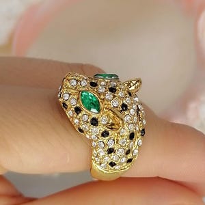 May include: A gold-toned ring shaped like a leopard head, featuring a pav&eacute; setting of clear white crystals and small black stones. The leopard's eyes are set with marquise-cut green stones, and the metal band has a polished gold finish.