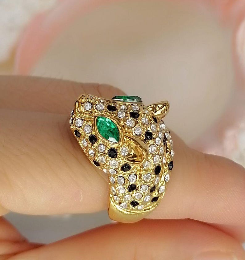 May include: A gold-toned ring shaped like a leopard head, featuring a pav&eacute; setting of clear white crystals and small black stones. The leopard's eyes are set with marquise-cut green stones, and the metal band has a polished gold finish.