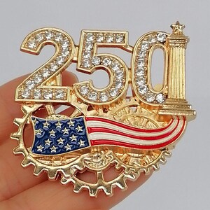 May include: Gold-toned brooch featuring the number "250" encrusted with clear rhinestones. The design includes a miniature American flag and a decorative column. The brooch is set against a backdrop of interlocking gears.